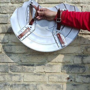 May include: A silver metal shield with brown leather straps and rivets. The shield is mounted on a brick wall. The shield is a historical replica, likely for costume or display.