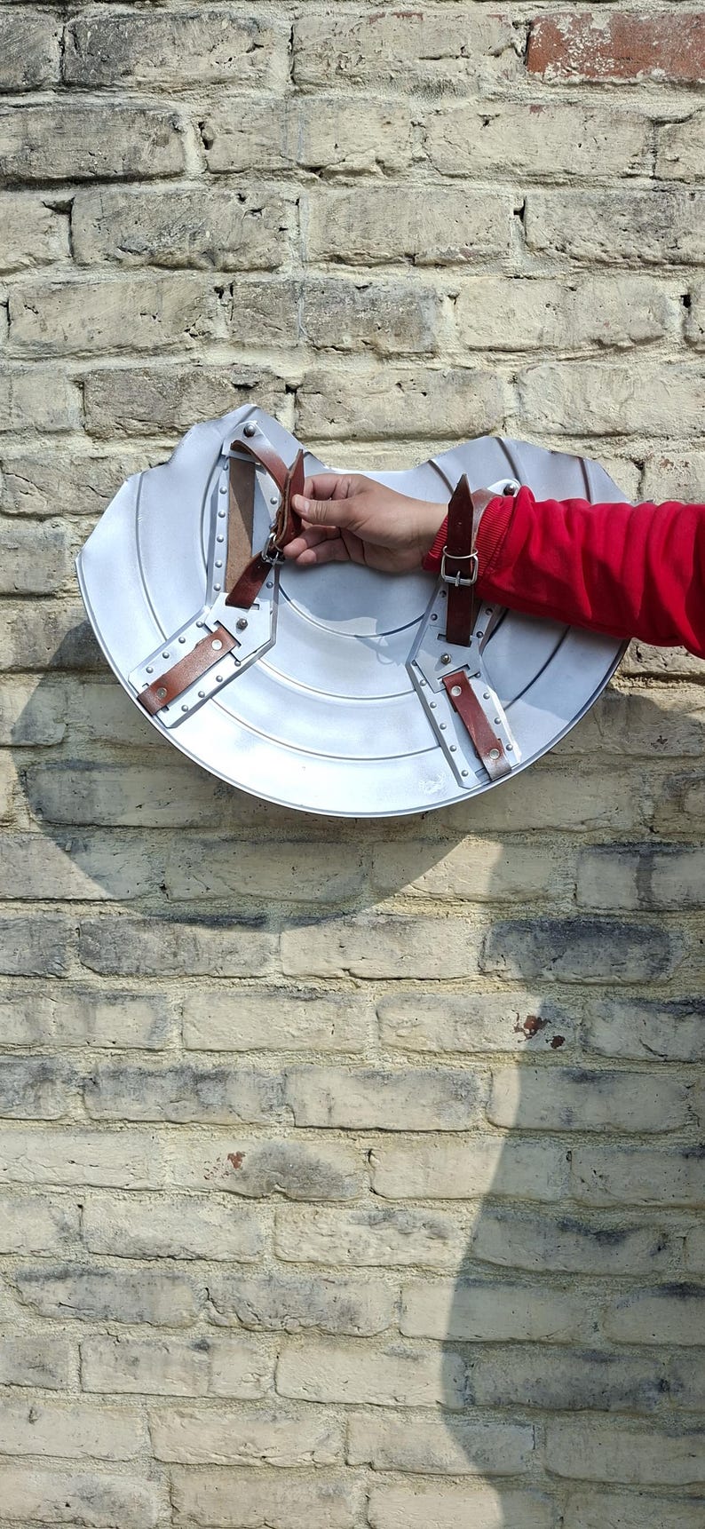 May include: A silver metal shield with brown leather straps and rivets. The shield is mounted on a brick wall. The shield is a historical replica, likely for costume or display.