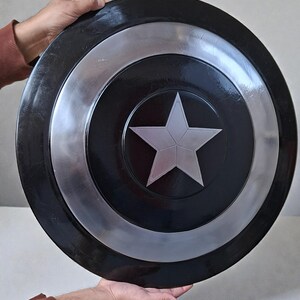 May include: A black and silver shield featuring a central silver star. The shield has a circular design with concentric rings of black and silver. The shield is held by two hands.
