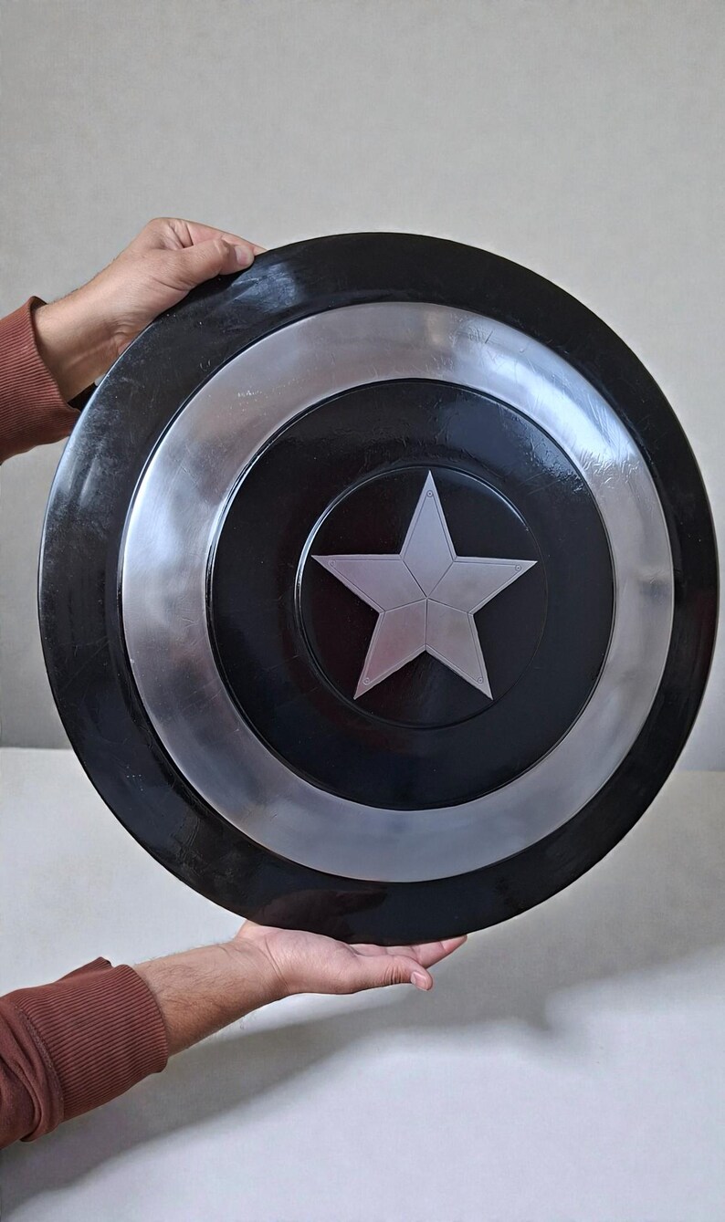May include: A black and silver shield featuring a central silver star. The shield has a circular design with concentric rings of black and silver. The shield is held by two hands.