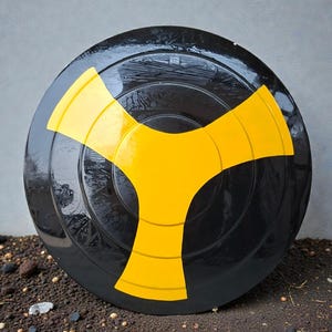 Puede incluir: Objeto circular negro brillante con un diseño amarillo brillante en forma de Y estilizada en el centro. El objeto se encuentra sobre un fondo neutro y descansa sobre un lecho de pequeñas piedras.