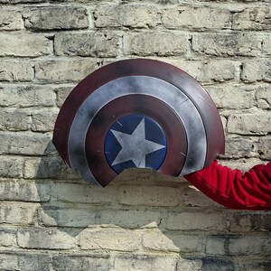 May include: A Captain America shield with a red, white, and blue color scheme. The shield features a central blue circle with a white star, surrounded by concentric rings of red and silver. The shield is held against a brick wall.