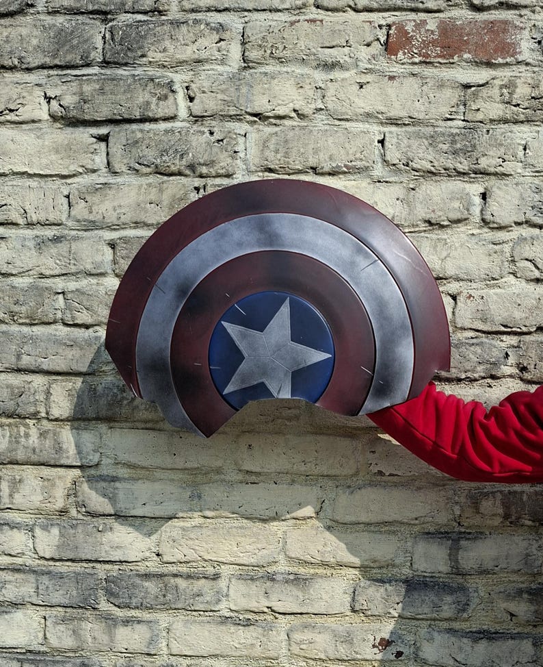 May include: A Captain America shield with a red, white, and blue color scheme. The shield features a central blue circle with a white star, surrounded by concentric rings of red and silver. The shield is held against a brick wall.