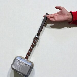 May include: A silver and gray hammer with a brown leather-wrapped handle and a leather strap. The hammer has a rectangular head and is held by a hand against a white background. The hammer is a replica of Thor's hammer.