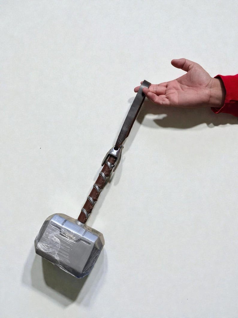 May include: A silver and gray hammer with a brown leather-wrapped handle and a leather strap. The hammer has a rectangular head and is held by a hand against a white background. The hammer is a replica of Thor's hammer.