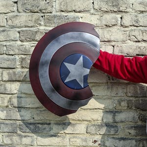 May include: A Captain America shield with a red, white, and blue color scheme. The shield has a star in the center and is held by a person wearing a red long-sleeve shirt. The shield is against a brick wall.
