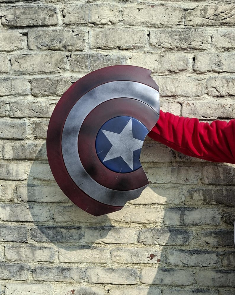 May include: A Captain America shield with a red, white, and blue color scheme. The shield has a star in the center and is held by a person wearing a red long-sleeve shirt. The shield is against a brick wall.