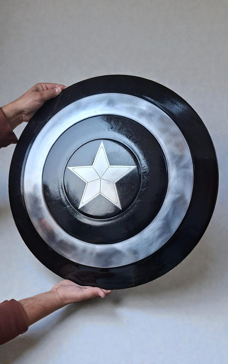 May include: A black and silver shield featuring a white star at its centre. The shield has a circular design with black and silver concentric rings. The star is a five-pointed design. The shield is held by two hands.