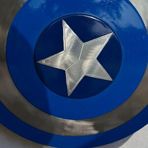 May include: A close-up of a shield with a blue, silver, and metallic design. The shield has a blue outer ring, a silver middle ring, and a blue center with a silver star. The shield is a replica of Captain America's shield.