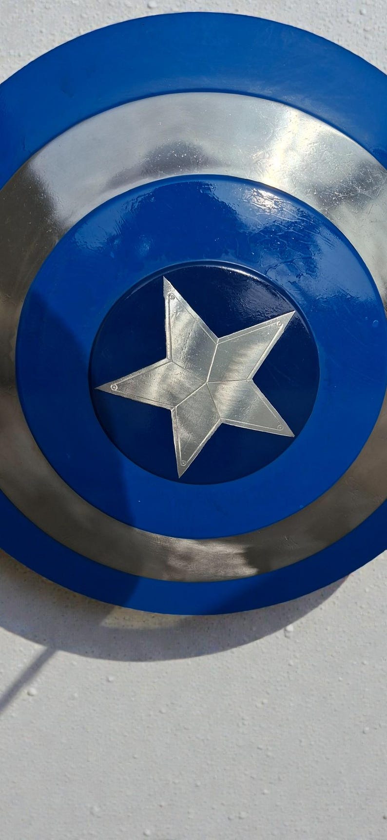 May include: A close-up of a shield with a blue, silver, and metallic design. The shield has a blue outer ring, a silver middle ring, and a blue center with a silver star. The shield is a replica of Captain America's shield.