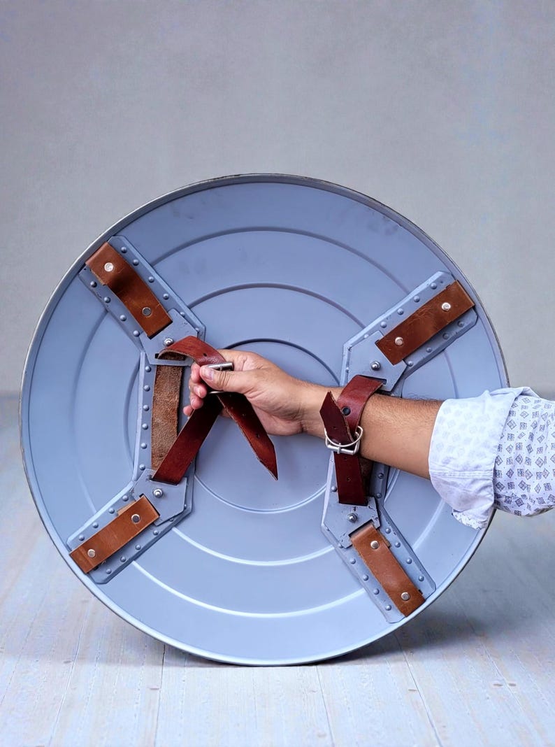 Puede incluir: Un escudo redondo azul claro con correas de cuero marr&oacute;n y detalles met&aacute;licos. El escudo tiene un asa y una correa para el brazo para un agarre seguro. El dise&ntilde;o recuerda a un escudo de superh&eacute;roe.
