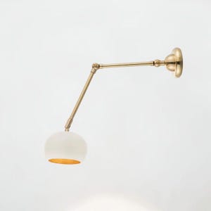 May include: A wall-mounted lamp featuring a brass-toned metal arm with two adjustable joints and a circular mounting plate. The lamp includes a white, dome-shaped shade with a gold-toned interior finish, designed to direct light downward.