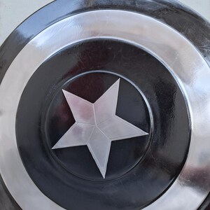 May include: A circular shield featuring a silver star at its centre, encircled by bands of silver, black, and silver. The shield has a shiny finish and seems to be made of metal. The design is reminiscent of a superhero's shield.