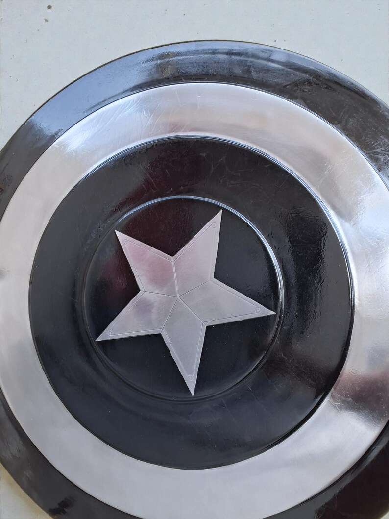 May include: A circular shield featuring a silver star at its centre, encircled by bands of silver, black, and silver. The shield has a shiny finish and seems to be made of metal. The design is reminiscent of a superhero's shield.