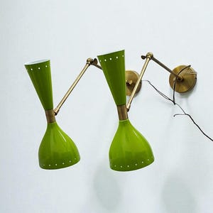 May include: Two green wall-mounted lamps with brass-colored adjustable arms and circular wall mounts. The lampshades are cone-shaped with white dotted accents. A black wire extends from one lamp.