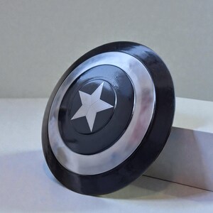 May include: A black and silver shield with a white star in the centre. The shield has a circular design with a silver border and a black centre. The shield is a prop or collectible.
