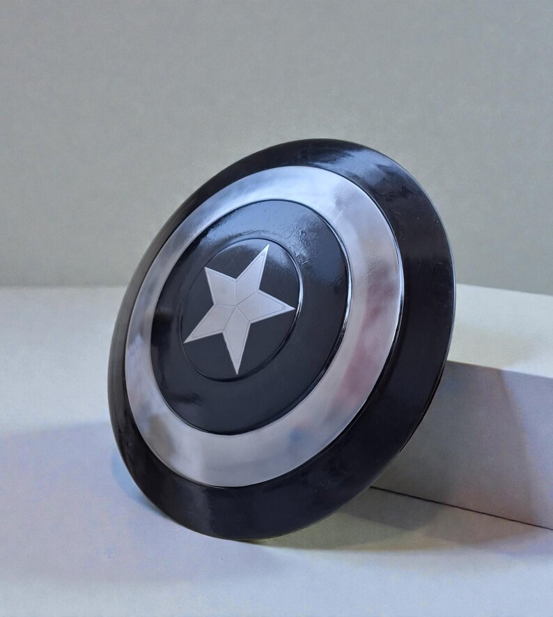 May include: A black and silver shield with a white star in the centre. The shield has a circular design with a silver border and a black centre. The shield is a prop or collectible.