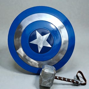 May include: A blue, silver, and white Captain America shield with a silver star in the center. A silver hammer with a brown handle and leather strap rests below the shield. The shield is round.