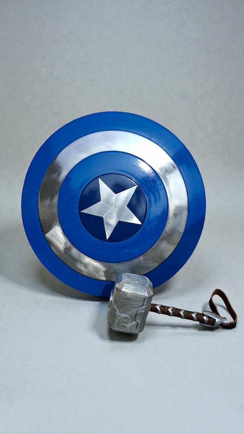 May include: A blue, silver, and white Captain America shield with a silver star in the center. A silver hammer with a brown handle and leather strap rests below the shield. The shield is round.