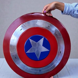 Puede incluir: Un escudo circular rojo, blanco y azul, con una estrella plateada en el centro. El escudo tiene un anillo plateado alrededor del centro. El escudo est&aacute; sostenido por dos manos.