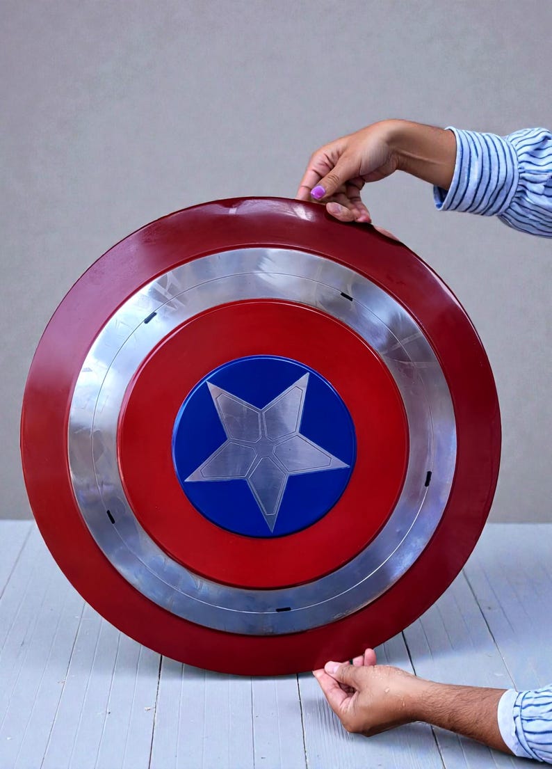 Puede incluir: Un escudo circular rojo, blanco y azul, con una estrella plateada en el centro. El escudo tiene un anillo plateado alrededor del centro. El escudo est&aacute; sostenido por dos manos.