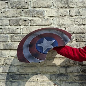 May include: A Captain America shield, with a blue star on a blue circle, surrounded by red and white stripes, is held against a brick wall. The shield is damaged, with a piece broken off. A red sleeve is visible.