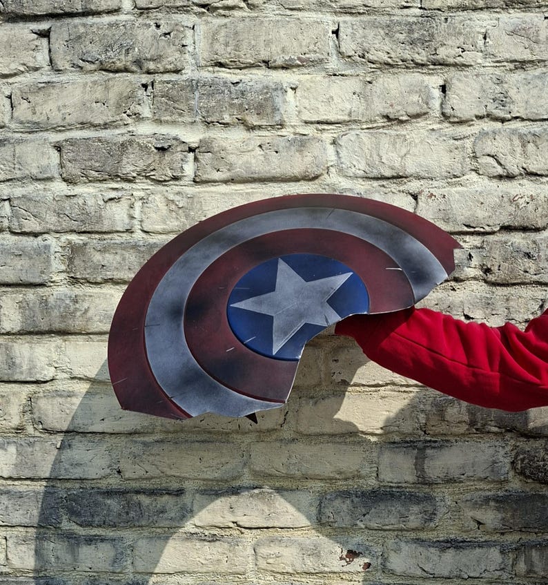 May include: A Captain America shield, with a blue star on a blue circle, surrounded by red and white stripes, is held against a brick wall. The shield is damaged, with a piece broken off. A red sleeve is visible.