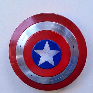 Puede incluir: Un escudo circular rojo, plateado y azul con un emblema de estrella blanca. El escudo tiene un anillo exterior rojo, un anillo central plateado y un centro rojo. Un c&iacute;rculo azul en el centro muestra una estrella blanca.