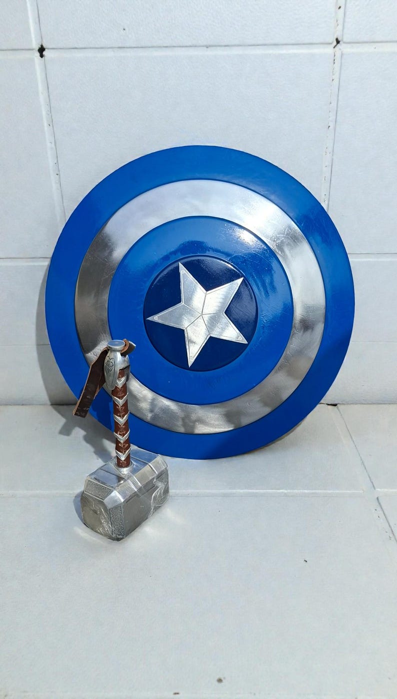 May include: A blue, silver, and white Captain America shield with a five-pointed star in the center. A silver and brown Thor's hammer is in front of the shield. The shield is approximately 24 inches in diameter.