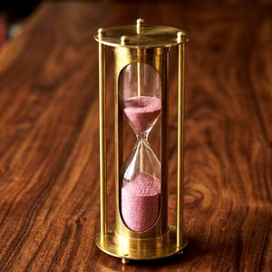 Könnte beinhalten: Eine Sanduhr mit einem Rahmen aus poliertem Messing, vertikalen Stützstangen sowie runden Platten an Ober- und Unterseite. Der zentrale Glaskolben enthält feinen, altrosa Sand, wobei sich ein Teil des Sandes in der unteren Kammer befindet.