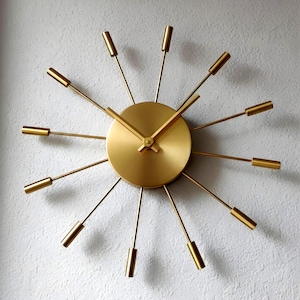 Puede incluir: Un reloj de pared dorado con diseño de rayos de sol, que presenta una esfera central circular con dos manecillas doradas a juego. Doce radios metálicos finos irradian desde el centro, cada uno con una tapa cilíndrica dorada, creando un diseño geométrico.