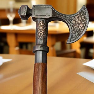 Op de afbeelding: Decoratieve wandelstok met een hamer- en bijlkopontwerp. De metalen kop heeft sierlijke bloempatronen en een donkere afwerking. De handgreep is gemaakt van gepolijst hout. De wandelstok is ontworpen voor decoratieve doeleinden.