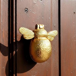 Può includere: Un battente per porta a forma di ape color oro, con dettagli strutturati e ali spiegate. Il battente è montato su una porta di legno marrone scuro. Il battente a forma di ape è alto circa 10 cm.