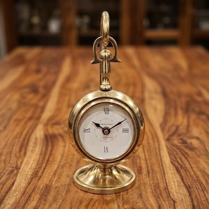 Puede incluir: Un reloj de mesa de latón con marco circular y base de pedestal. La esfera blanca presenta números romanos negros, manecillas negras y el texto 4 BOND STREET LONDON. Un asa vertical de latón se extiende desde la parte superior del reloj.
