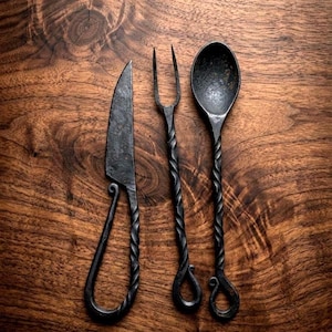 May include: Hand-forged black metal cutlery set including a knife, fork, and spoon. The knife has a curved handle, the fork has two tines, and the spoon has a rounded bowl. The handles are twisted, and the set is displayed on a wooden surface.