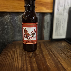 May include: A dark brown glass bottle of Big Js Back Porch Sauce, with a red and white label featuring a cartoon figure. The bottle is 12 fl oz (360 ml).