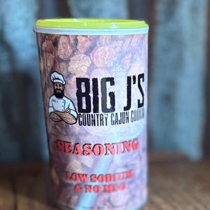Peut inclure: Un récipient cylindrique de l'assaisonnement de cuisine cajun de Big J. L'étiquette présente une illustration de chef et le texte "SEASONING" en rouge. Le récipient a un couvercle jaune et un fond de ce qui semble être des saucisses séchées.