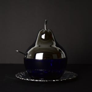 May include: A pear-shaped serving dish with a silver-toned lid and a dark blue base, accompanied by a small spoon. It rests on a decorative silver plate, set against a dark backdrop.