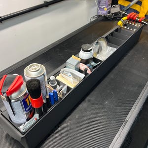 Może przedstawiać: Czarny, prostokątny organizer z wieloma przegródkami, wypełniony różnymi narzędziami i materiałami. Zawiera puszki ze sprayem, pędzle, butelki i małe pojemniki. Organizer stoi na teksturowanej, czarnej powierzchni.