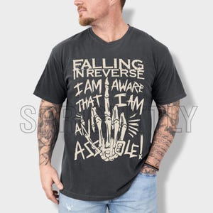 May include: Charcoal grey t-shirt featuring the text "FALLING IN REVERSE I AM AWARE THAT I AM AN ASSHOLE!" in a distressed, off-white font. The graphic includes a skeletal hand giving the middle finger.