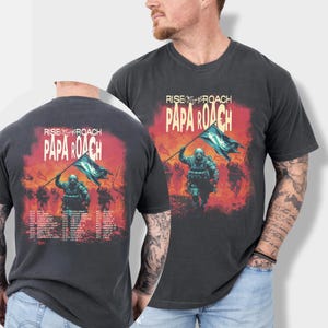 May include: Dark gray t-shirt featuring the band Papa Roach's "Rise of the Roach" design. The graphic shows a soldier holding a flag, with a fiery background. The back of the shirt includes a tour date list.