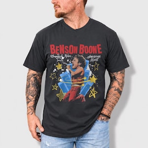 May include: Dark gray t-shirt featuring a graphic of Benson Boone performing on stage. The design includes the text "Benson Boone" in red, with "Wanted Man Tour" and "American Heart" details. The image has blue and yellow star accents.