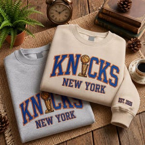 May include: Two crewneck sweatshirts, one in light grey and one in beige, each featuring the text KNICKS NEW YORK in blue and orange collegiate lettering with a basketball trophy graphic. The beige sleeve includes the embroidered text EST. 2026.