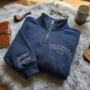 Peut inclure: Un sweat-shirt bleu marine à fermeture éclair quart de col avec le mot "GRANDPA" brodé en blanc, ainsi que "EST 2025". La manche gauche porte les noms "James Sophia" brodés en blanc. Le sweat-shirt est sur un tapis gris duveteux.