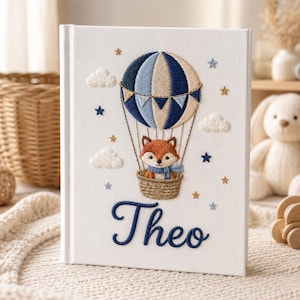 Könnte beinhalten: Ein weißer Buchumschlag mit einem gestickten Design, das einen Fuchs in einem Heißluftballon zeigt. Der Ballon ist blau und beige, mit Wolken und Sternen. Der Name "Theo" ist darunter in Blau gestickt.