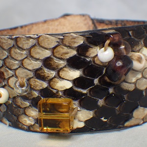 May include: A brown leather cuff bracelet with a snakeskin pattern and various colored beads. The bracelet has a large amber-colored square bead.