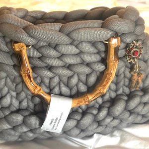 May include: A gray, chunky-knit handbag with bamboo handles and a decorative charm. The bag features a braided top handle and a rectangular shape. A tag is attached to the handle. The charm has red and silver accents.
