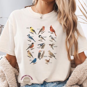 May include: Ivory-colored t-shirt featuring a colorful illustration of various bird species, each labeled with its name. The design includes birds like the American Goldfinch, Blue Jay, and Northern Cardinal. The shirt has the text "COMFORT COLORS" and "IVORY".