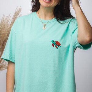 May include: A light mint green short-sleeve t-shirt featuring a small embroidered sea turtle on the left chest. The turtle has a bright red shell with dark red spots, a teal body, and large eyes. The shirt has a crew neckline and a relaxed fit.