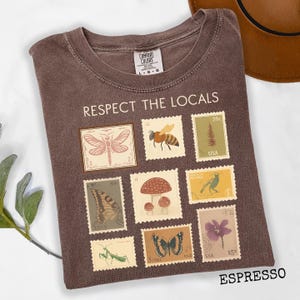 May include: Espresso-colored t-shirt with the text "RESPECT THE LOCALS" and a collection of illustrated stamps featuring insects, plants, and a bird. The shirt is a Comfort Colors brand.